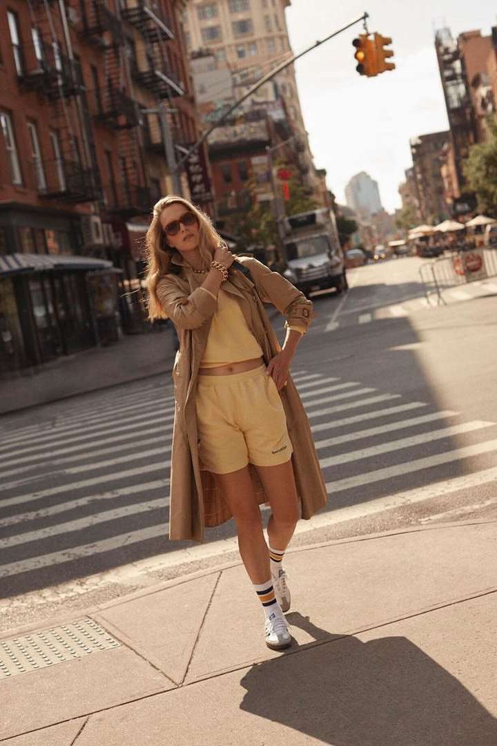 Woman in tan coat and yellow shorts stands on a crosswalk in an urban setting with brick buildings on a sunny day.