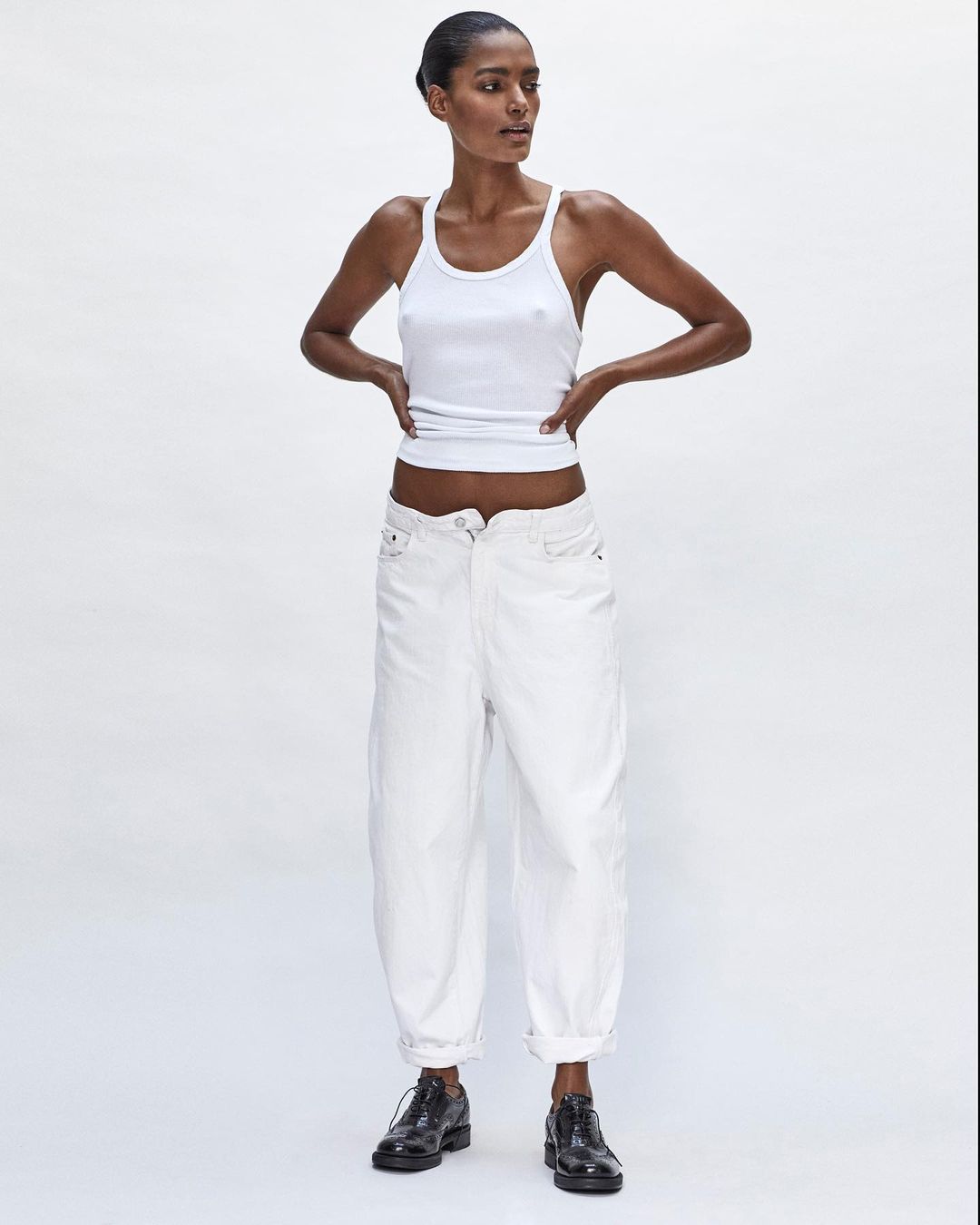Full-length shot of a Black female model posing in a white ribbed tank top, white balloon pants, and black dress shoes against a white backdrop.