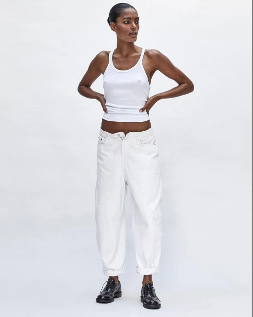 Full-length shot of a Black female model posing in a white ribbed tank top, white balloon pants, and black dress shoes against a white backdrop.