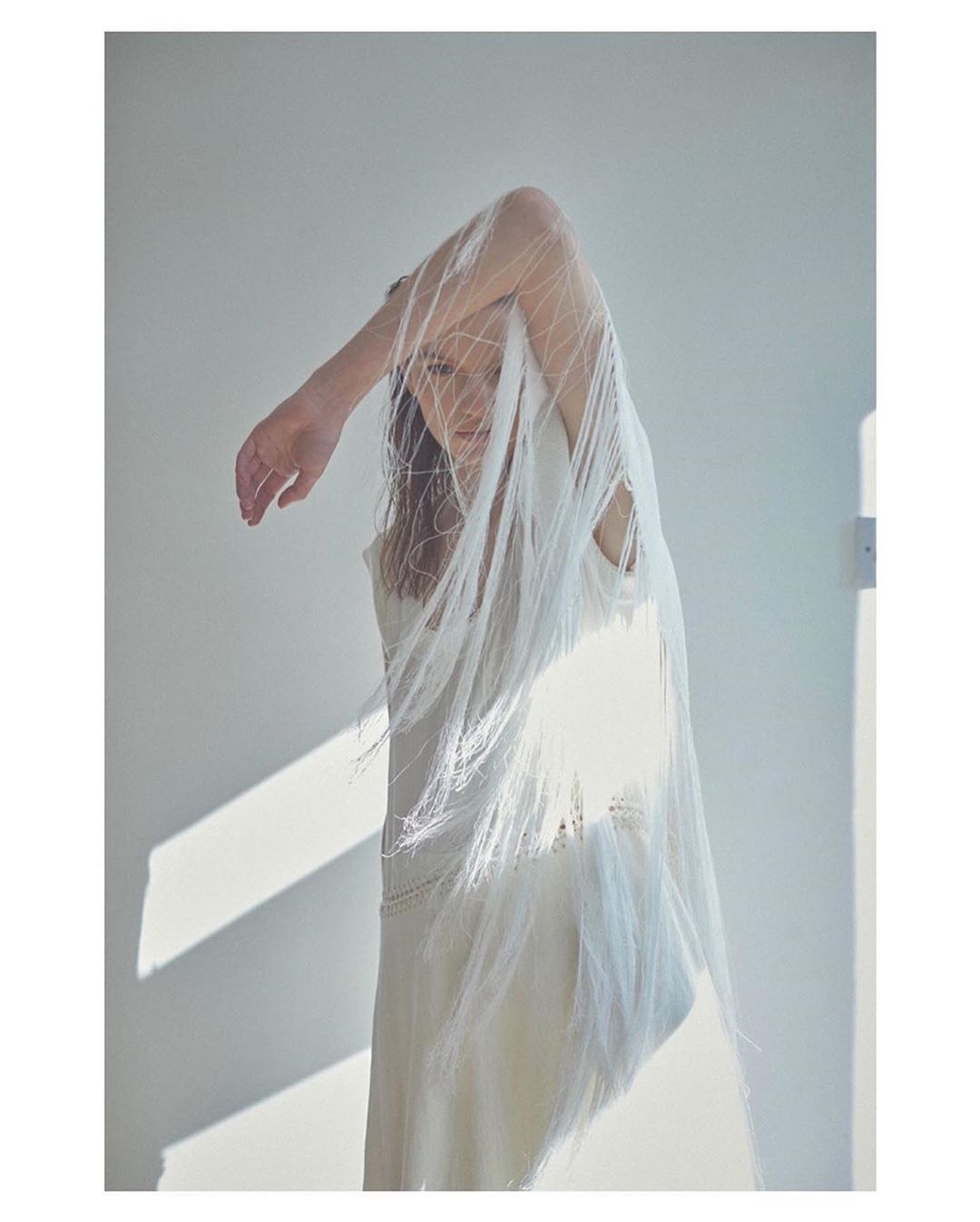 An adult female stands indoors draped in a white fringed fabric. The woman poses with an arm raised against a white wall illuminated by sunlight.