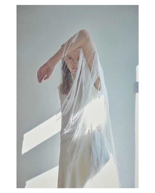An adult female stands indoors draped in a white fringed fabric. The woman poses with an arm raised against a white wall illuminated by sunlight.