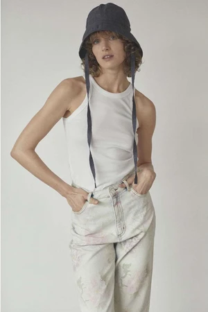Model with curly hair wearing a dark bucket hat, white tank top, and light patterned jeans against a plain white background.