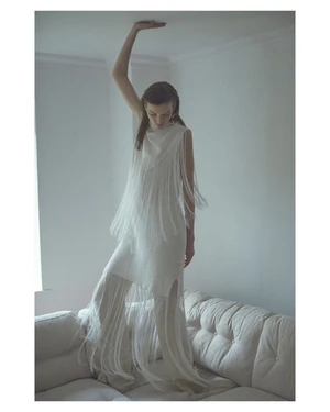 Model wearing white fringed dress standing on couch with hand touching ceiling in New York by Chloe Mallett