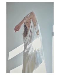 Model in white dress with sheer veil draped over raised arm in natural light by Chloe Mallett
