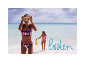 Smiling woman wearing a striped bikini adjusting a snorkel mask at the beach with a child walking behind her in the surf.