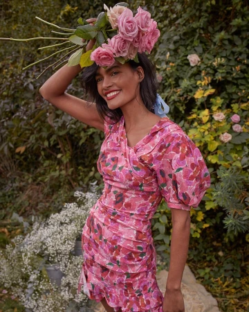 Smiling woman in pink floral dress holding a bouquet of pink roses above her head in a garden by Chloe Mallett