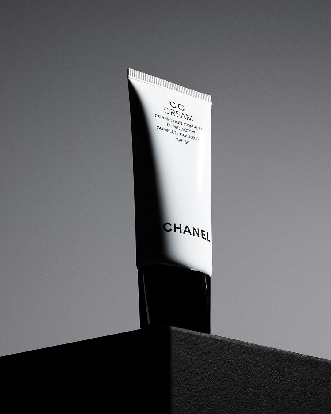 Still life studio shot of a white Chanel CC cream tube with black lettering, against a grey background on a black platform.