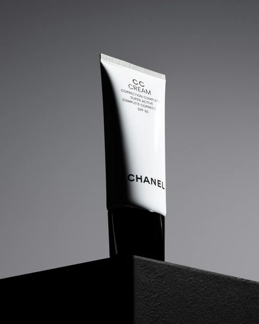 Still life studio shot of a white Chanel CC cream tube with black lettering, against a grey background on a black platform.