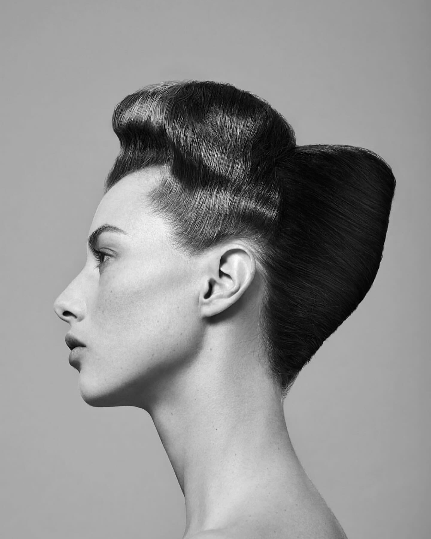 Monochrome portrait of a woman in profile, showcasing an elaborate vintage hairstyle against a simple grey background.