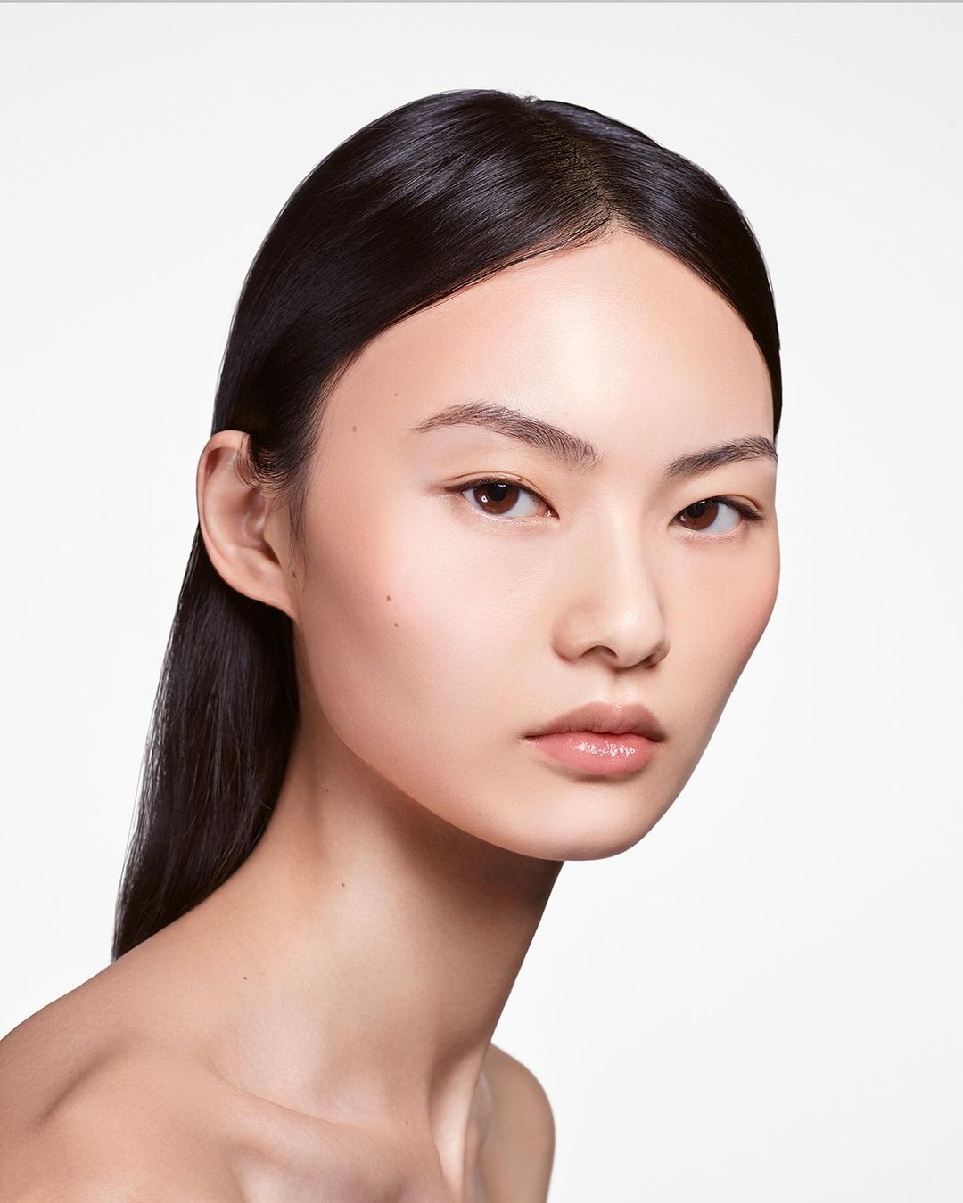 Close-up beauty shot: young Asian woman with bare shoulders, slicked black hair, and flawless makeup against a bright white background.