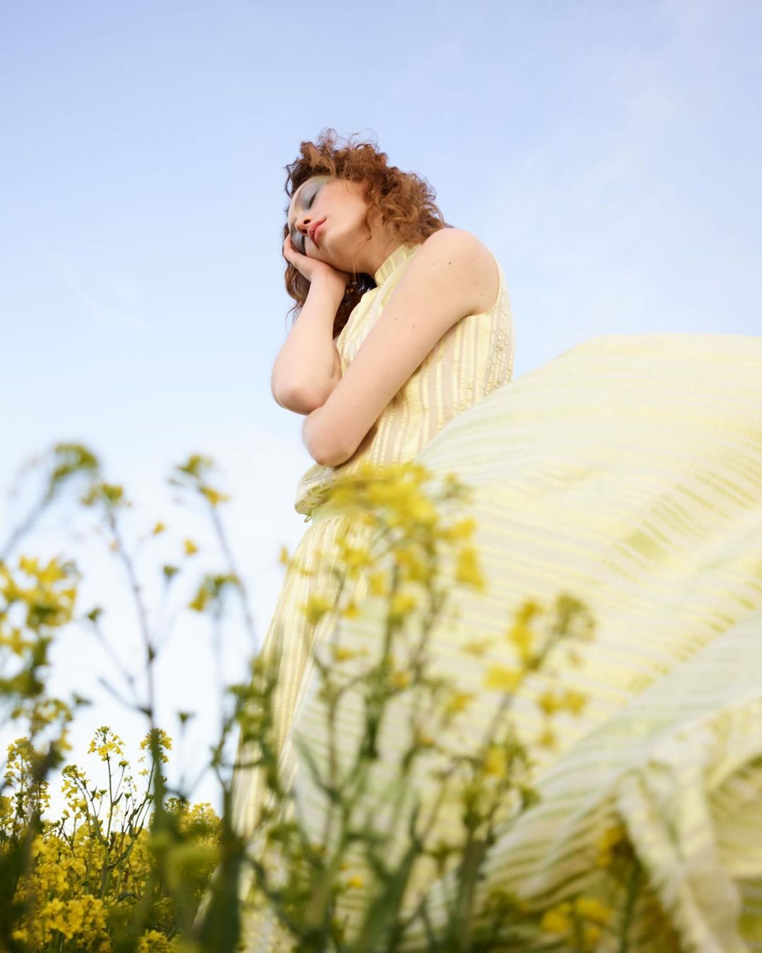 Young woman in yellow dress rests in a field of yellow flowers against a light blue sky in an outdoor fashion portrait.