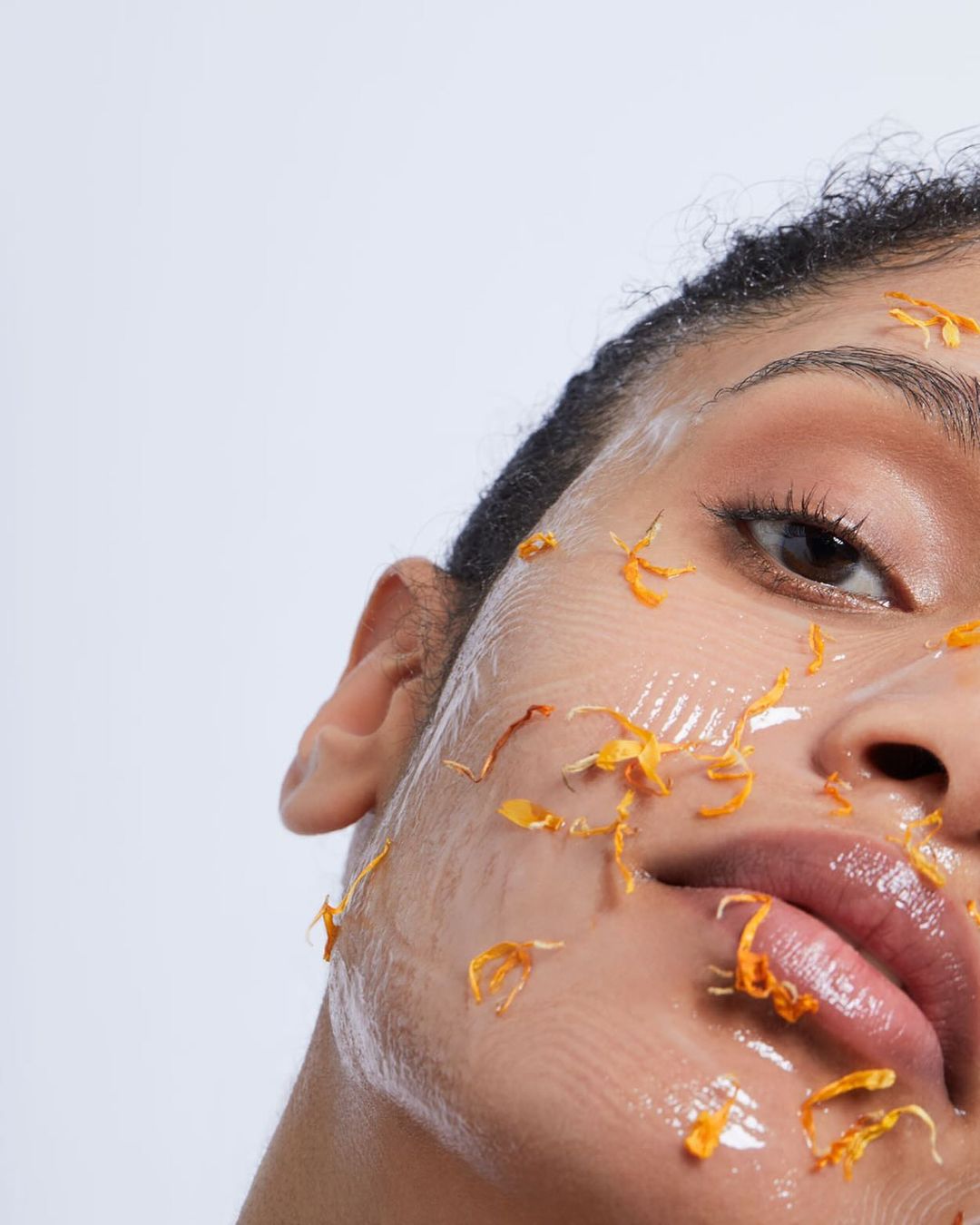 Close-up of a woman's face covered in a clear gel and orange petals. One eye is visible with a light skin tone on a white background.
