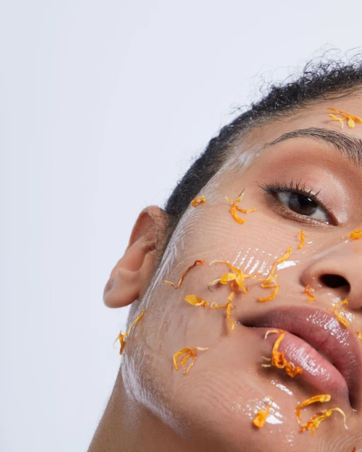 Close-up of a woman's face covered in a clear gel and orange petals. One eye is visible with a light skin tone on a white background.