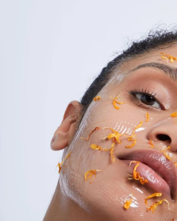 Close-up of a woman's face covered in a clear gel and orange petals. One eye is visible with a light skin tone on a white background.