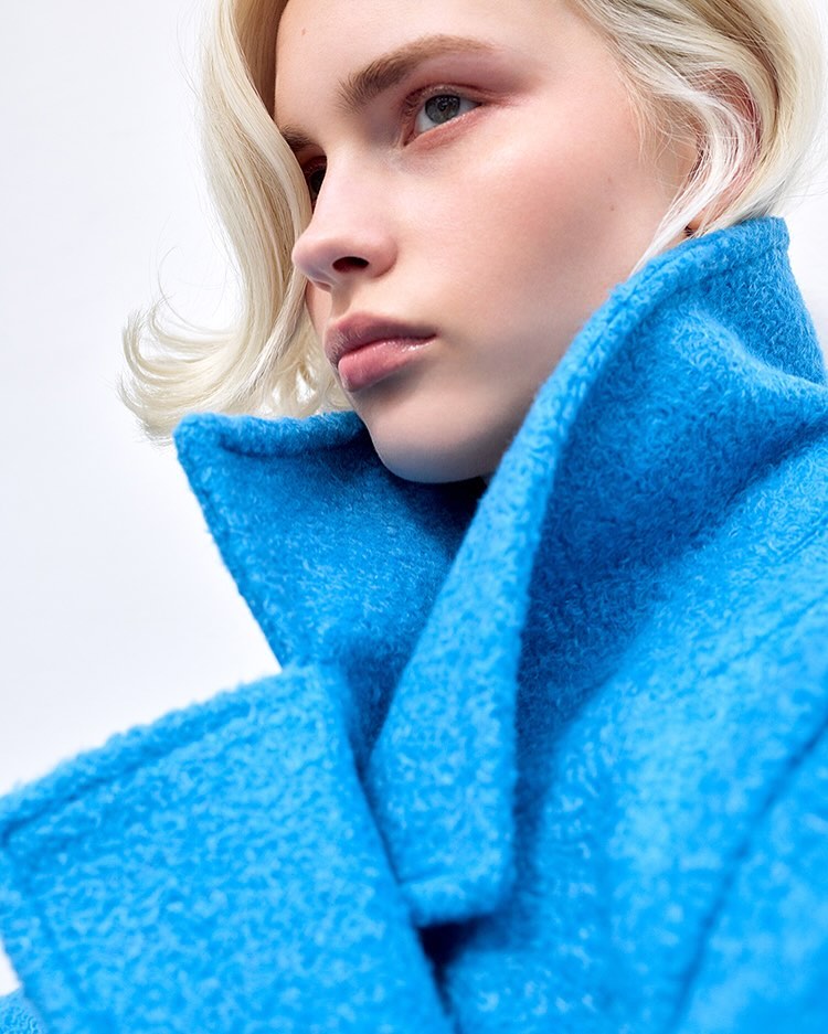 Close-up of a woman with platinum blonde hair wearing a textured turquoise coat against a plain white background in a fashion shot.