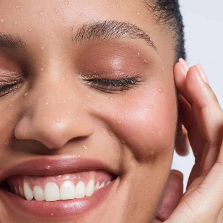 Close-up of smiling woman's face with water droplets on her skin, highlighting skincare and beauty, set against a clean white background.