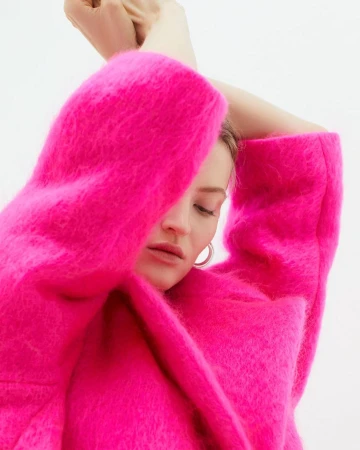 Model in vibrant hot pink fuzzy mohair coat with arms raised against a clean white background