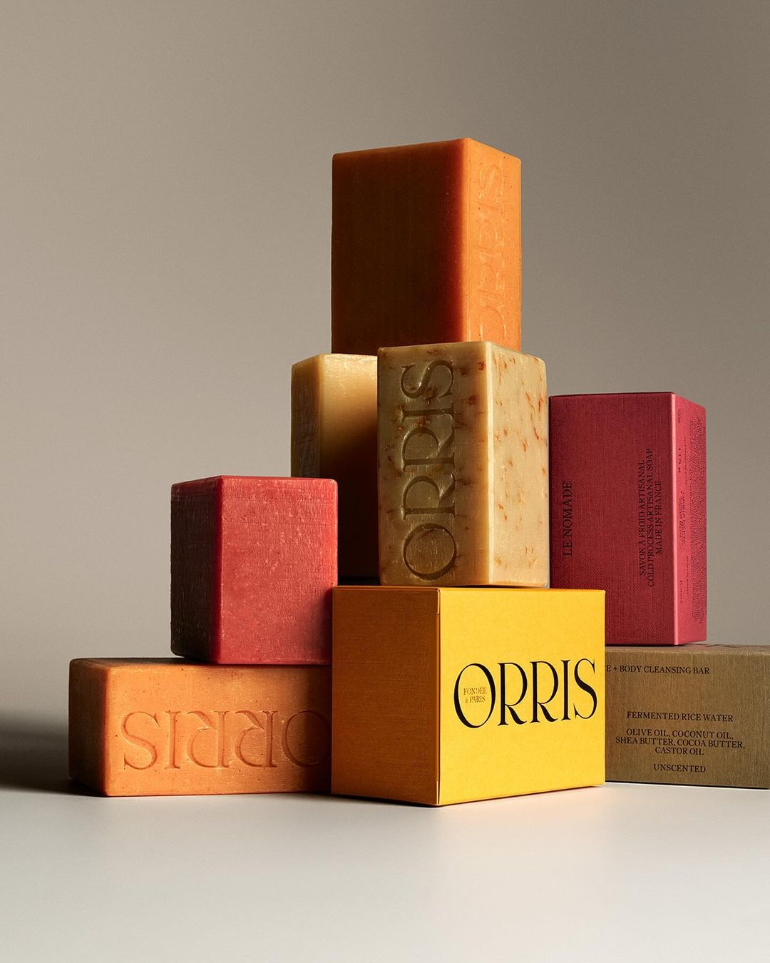 Stacked assortment of Orris scented soap bars in orange, yellow and red hues, creatively arranged on a light neutral surface.