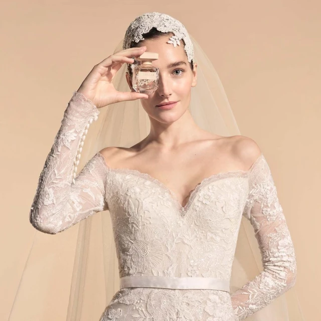 Portrait of a woman in a wedding dress and veil holding a perfume bottle in front of her face against a light brown background.