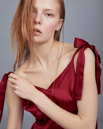 Close-up portrait of a female model in a red silk camisole with shoulder bows against a plain grey backdrop.