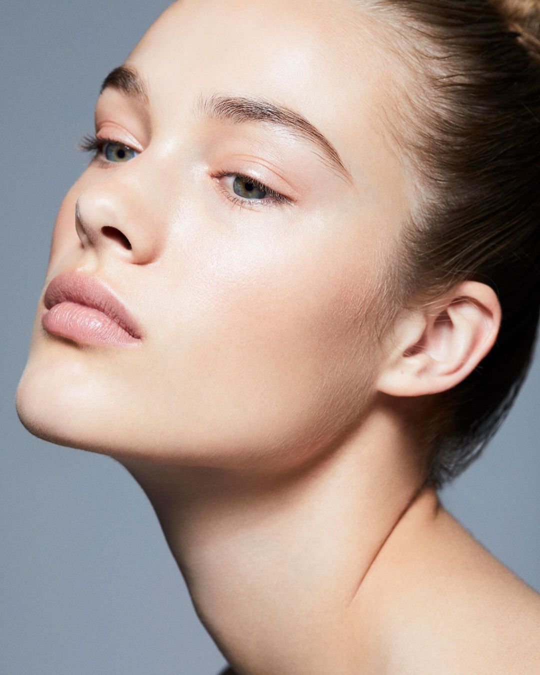 Close-up studio shot of a young woman with fair skin, natural makeup, and slicked-back hair against a solid grey background.