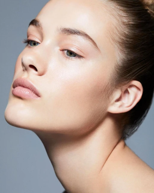Close-up studio shot of a young woman with fair skin, natural makeup, and slicked-back hair against a solid grey background.