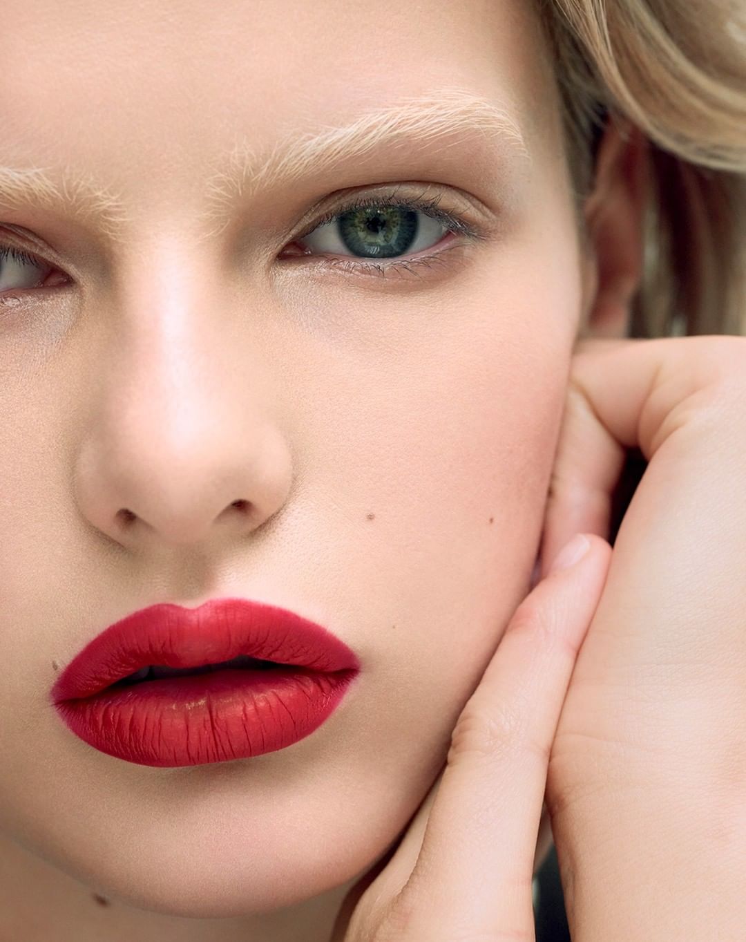 Close-up beauty portrait of a woman with vibrant red lipstick, blonde eyebrows, and a hand resting on her cheek.