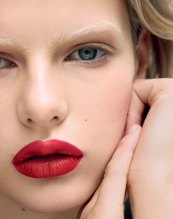 Close-up beauty portrait of a woman with vibrant red lipstick, blonde eyebrows, and a hand resting on her cheek.