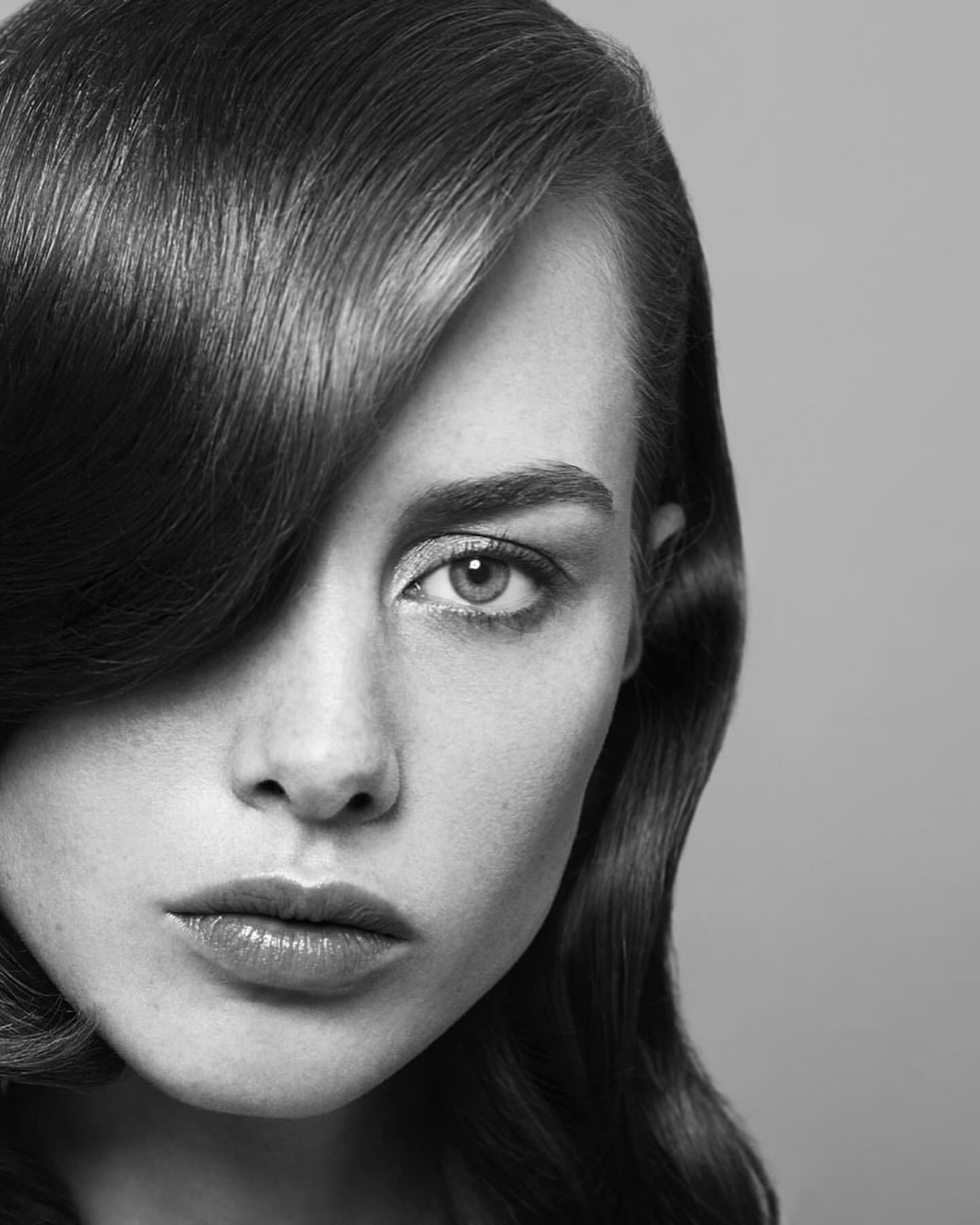 Monochrome portrait of a young fair-skinned woman with dark hair partially covering her face, gazing directly at the camera.