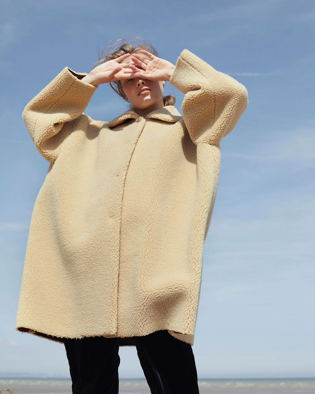 Woman shields eyes with hands in beige fleece coat against blue sky. Beach and ocean horizon in background. Fashion editorial style.