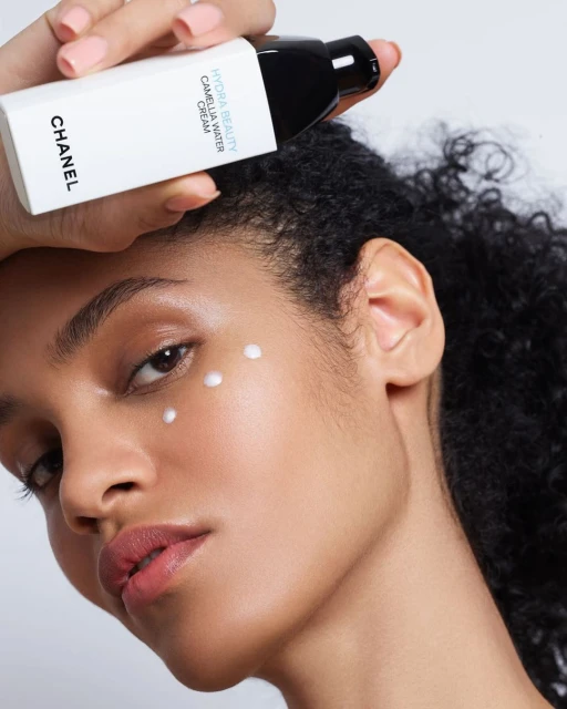 Close-up of a woman with skincare product applied under her eye, holding a bottle of Chanel Hydra Beauty Camellia Water Cream.