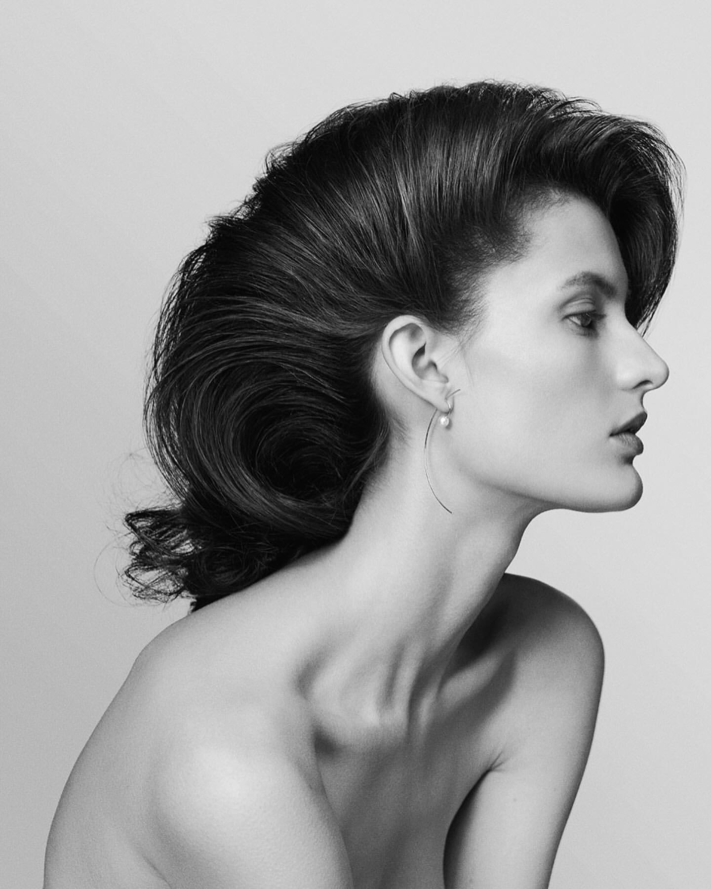 Striking black and white studio portrait of a poised adult female with voluminous dark hair, bare shoulders, and delicate pearl drop earrings.