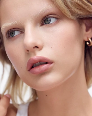 Close-up portrait of a blonde adult woman with light skin, wearing small gold hoop earrings. Beauty and fashion photography.