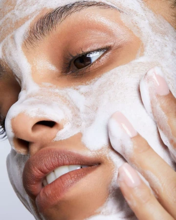 Close-up portrait of a woman with an even skin tone, light pink manicure, and white foamy cleanser applied to her face and hand.