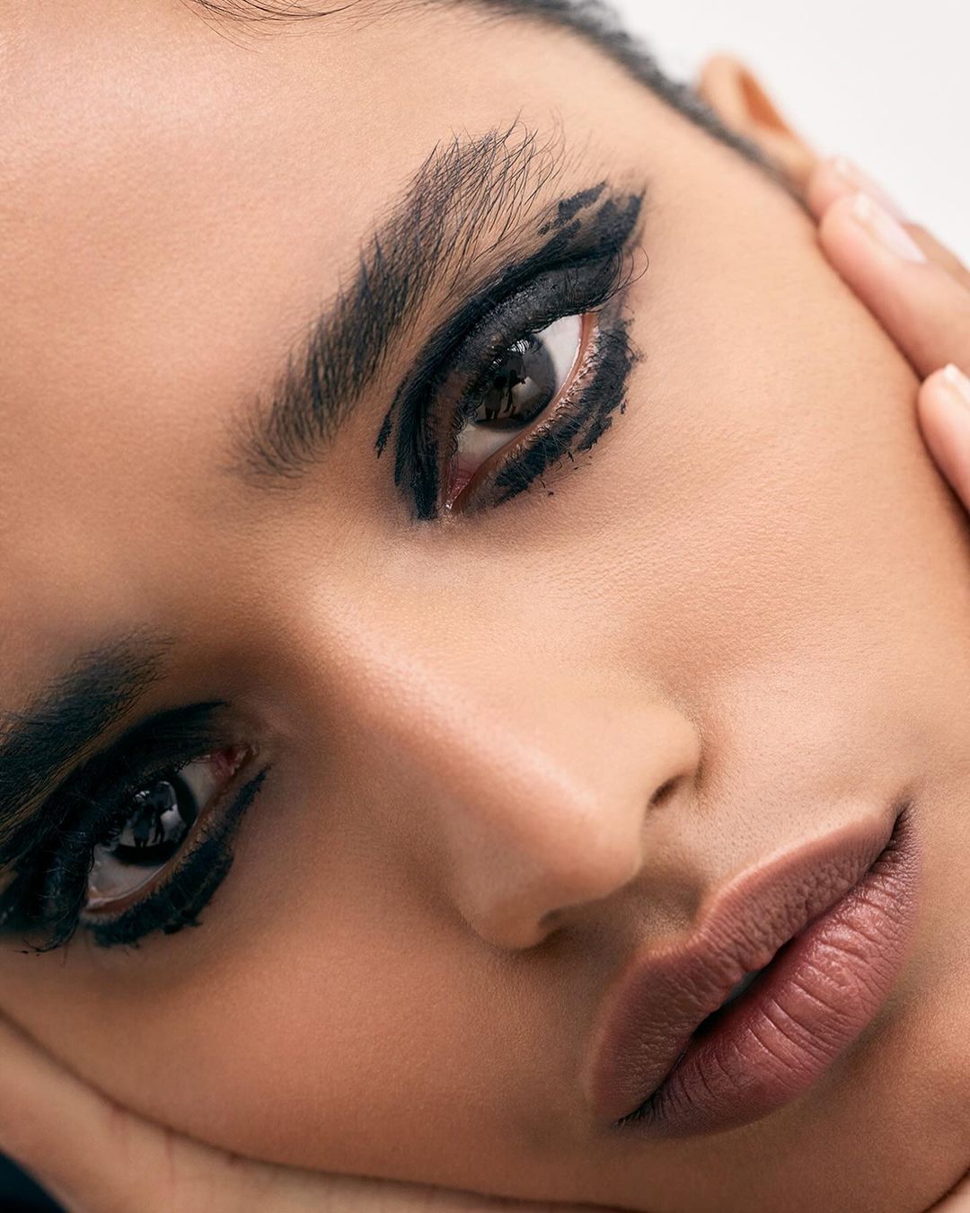 Close-up portrait of a woman with dark hair and dramatic black eyeshadow, gazing directly at the camera with her hands near her face.