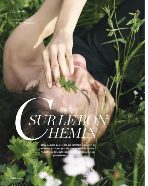 A woman is lying in green grass with a leaf on her face. Elle Beauty magazine cover with title 'Sur Le Bon Chemin'.