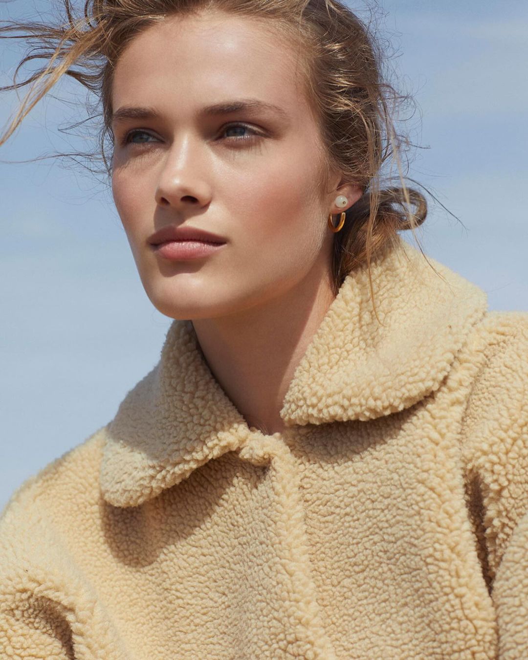 Close-up of a fair-skinned adult female model in a cream-colored fleece jacket against a light blue sky, wearing gold hoop earrings.