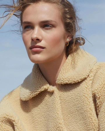 Close-up of a fair-skinned adult female model in a cream-colored fleece jacket against a light blue sky, wearing gold hoop earrings.
