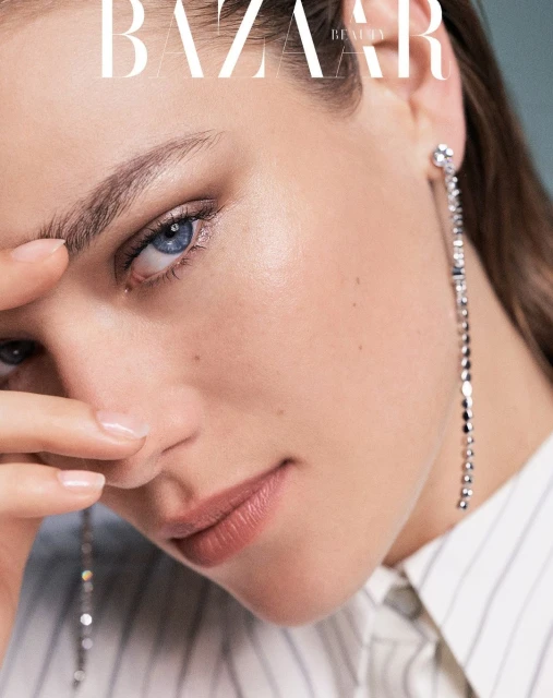 Close-up of a fair-skinned woman with blue eyes, wearing a white button-down shirt and long crystal earrings, and Bazaar Beauty text.