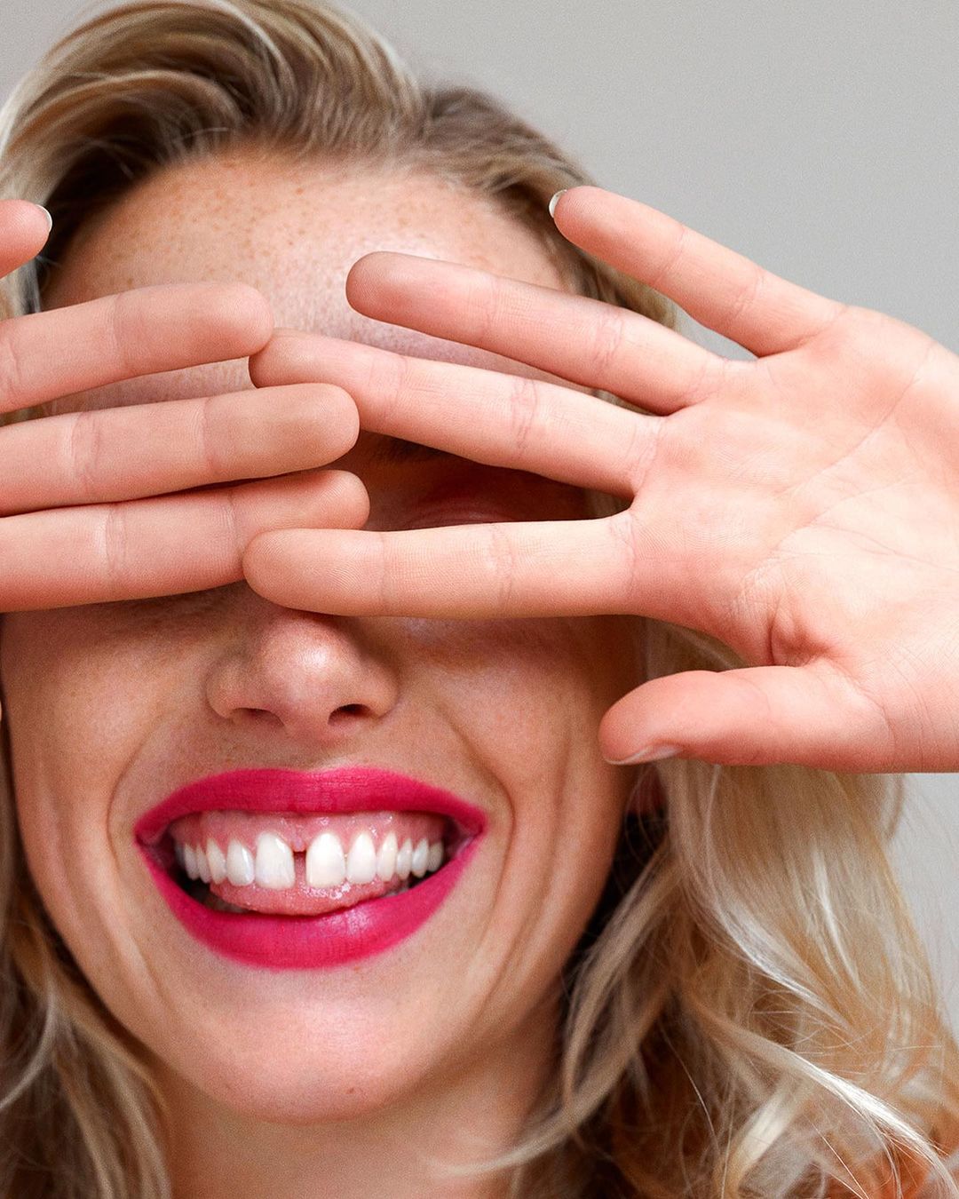 Close-up portrait of a blonde woman smiling widely with pink lipstick, holding both hands up to cover her eyes.
