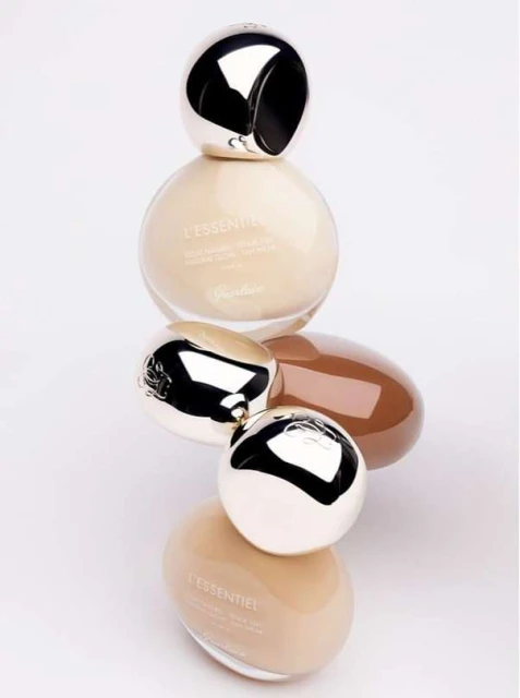 Stacked Guerlain L'Essentiel foundation bottles in various shades, featuring reflective silver caps, against a neutral background.