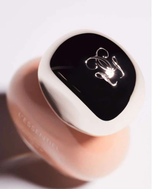 Close-up of Guerlain L'Essentiel foundation bottle with a glossy black and white cap and peach-toned container against a white background.
