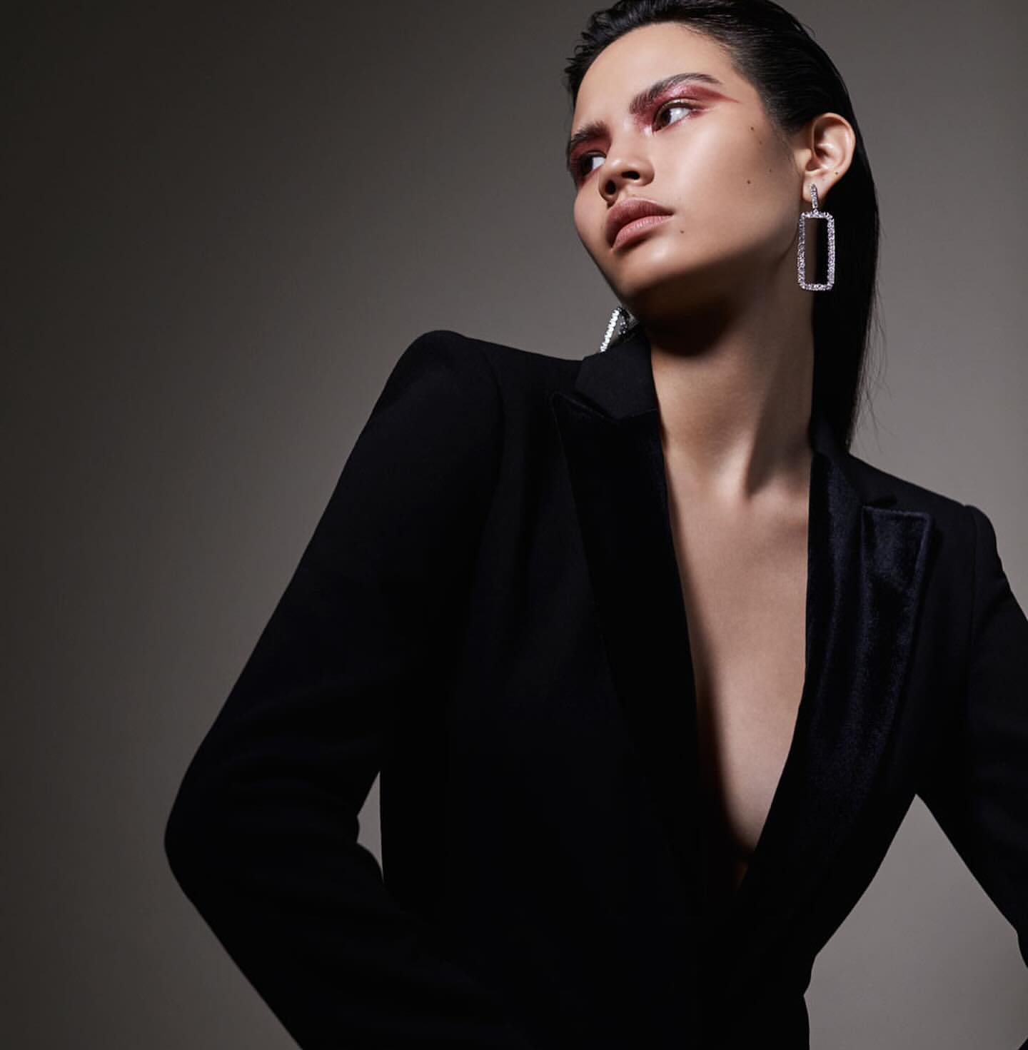 Portrait of a stylish brunette woman with a black blazer and statement earrings against a gray backdrop in a beauty and fashion shoot.
