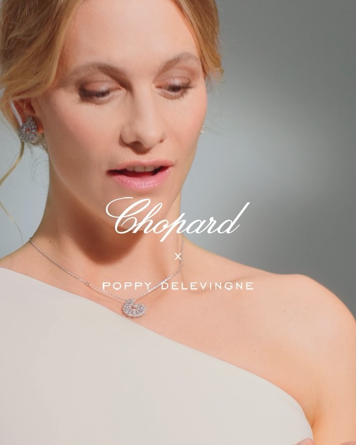 A fair-skinned woman with blonde hair wears a white, one-shoulder dress and jewelry in a studio shot on a grey background.