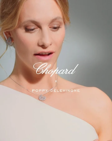 A fair-skinned woman with blonde hair wears a white, one-shoulder dress and jewelry in a studio shot on a grey background.