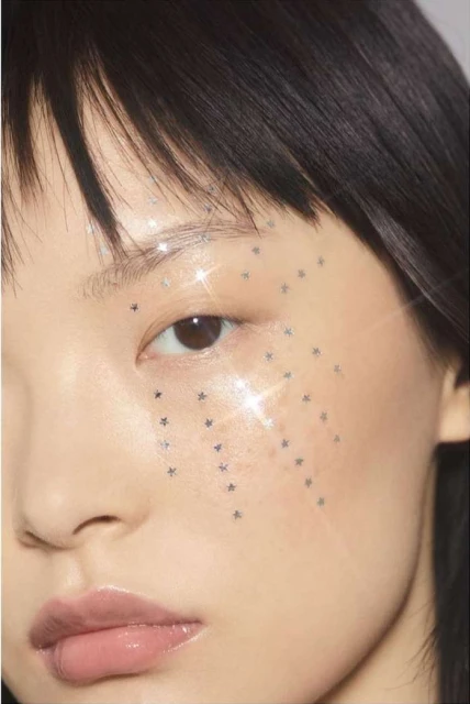Close-up portrait of an East Asian woman with dark hair and a cascade of star-shaped glitter makeup on her cheek, creating a sparkling effect.