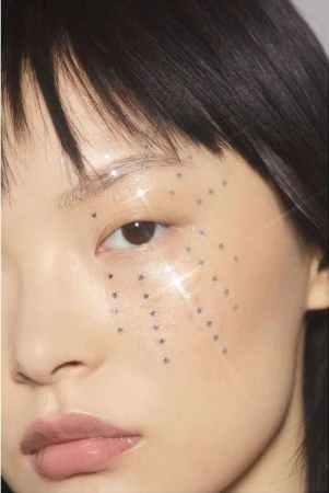 Close-up portrait of an East Asian woman with dark hair and a cascade of star-shaped glitter makeup on her cheek, creating a sparkling effect.