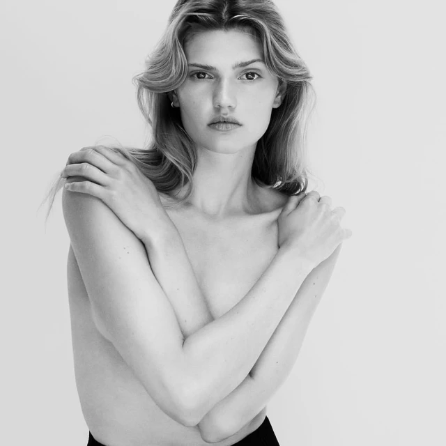 Black and white portrait of a fair-skinned adult female with shoulder-length wavy blonde hair, arms crossed, and a light background.