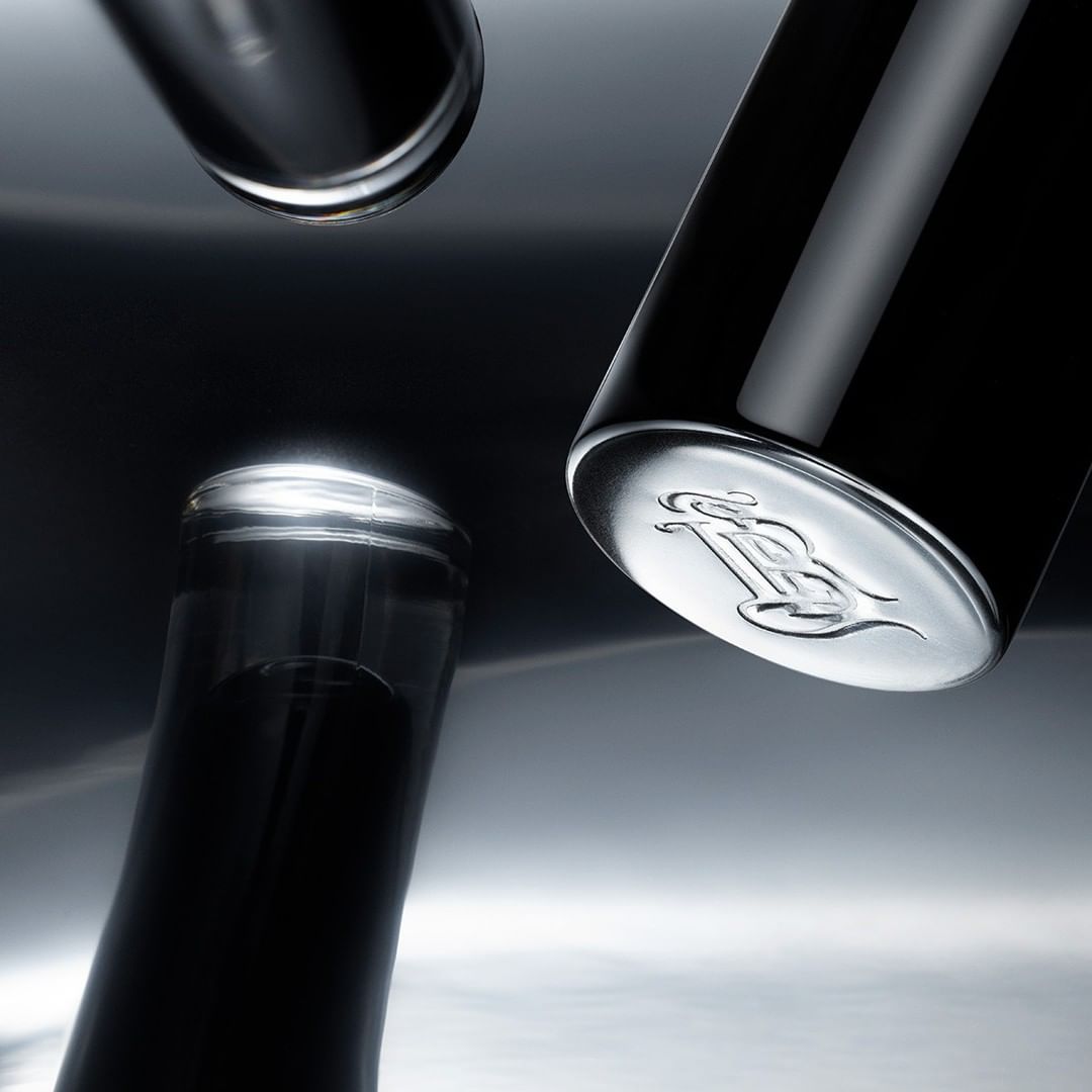 Close-up of three dark-colored bottles. One bottle shows an emblem at the bottom, set against a gradient grey background with reflections.
