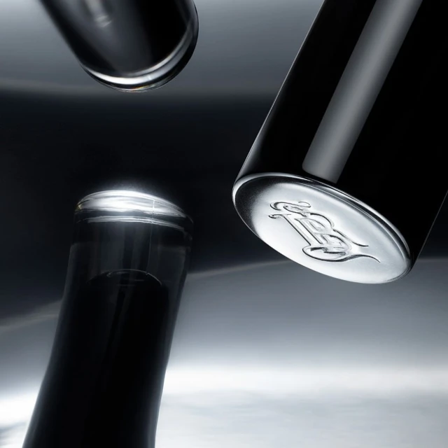 Close-up of three dark-colored bottles. One bottle shows an emblem at the bottom, set against a gradient grey background with reflections.
